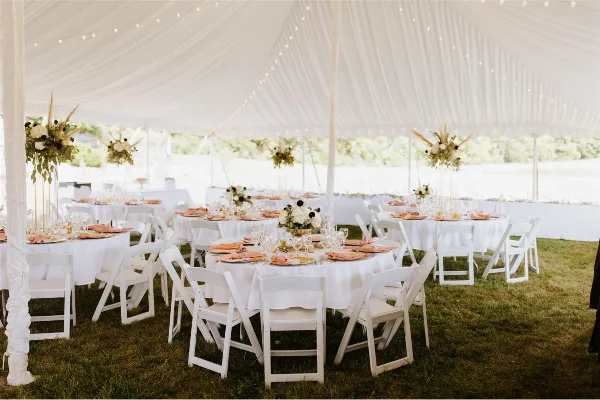 A wedding reception setup featuring white tables, chairs, and linens under a large white tent.