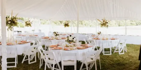 A wedding reception setup featuring white tables, chairs, and linens under a large white tent.