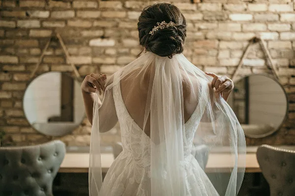 A bride from behind wearing a white lace wedding dress and veil in a room with a brick wall and mirrors.