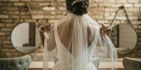 A bride from behind wearing a white lace wedding dress and veil in a room with a brick wall and mirrors.