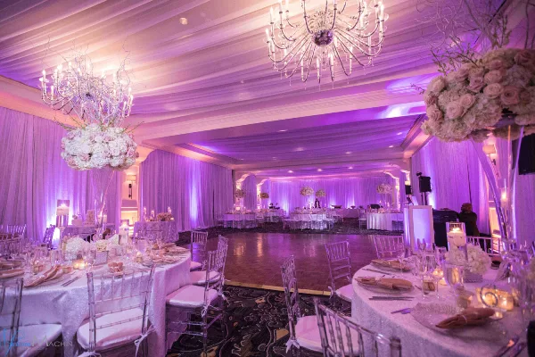 A luxurious wedding reception hall featuring purple ambient lighting, crystal chandeliers, and round tables set with white linens and tall floral arrangements.