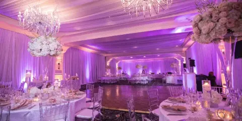 A luxurious wedding reception hall featuring purple ambient lighting, crystal chandeliers, and round tables set with white linens and tall floral arrangements.