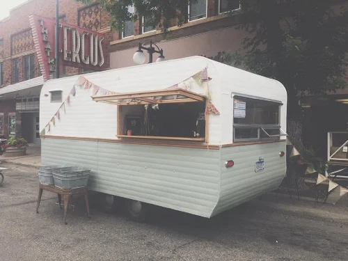 A vintage white camper trailer parked on a street, set up with a serving window and bunting.