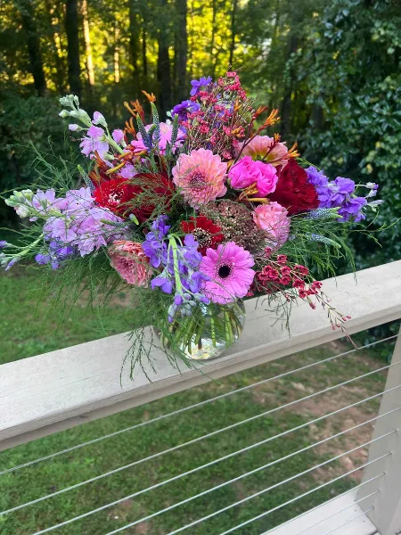 A colorful floral arrangement featuring roses, daisies, and gladioli in a glass vase, placed on a wooden deck railing.