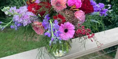 A colorful floral arrangement featuring roses, daisies, and gladioli in a glass vase, placed on a wooden deck railing.