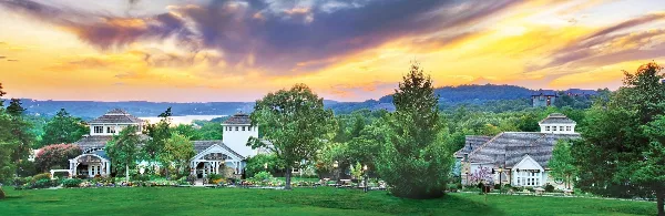 Wide shot of the Big Cedar Lodge resort buildings and green lawns at sunset with a lake and hills in the background.