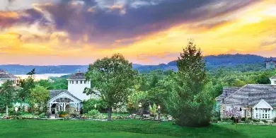 Wide shot of the Big Cedar Lodge resort buildings and green lawns at sunset with a lake and hills in the background.