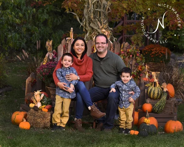 A professional outdoor family portrait of a couple and two young boys sitting on a bench surrounded by autumn decorations like pumpkins and corn stalks.