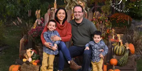 A professional outdoor family portrait of a couple and two young boys sitting on a bench surrounded by autumn decorations like pumpkins and corn stalks.