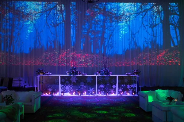 A decorated event venue featuring a floral head table, white sofas, and a large backdrop with a projected forest scene.