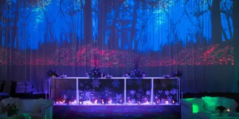 A decorated event venue featuring a floral head table, white sofas, and a large backdrop with a projected forest scene.