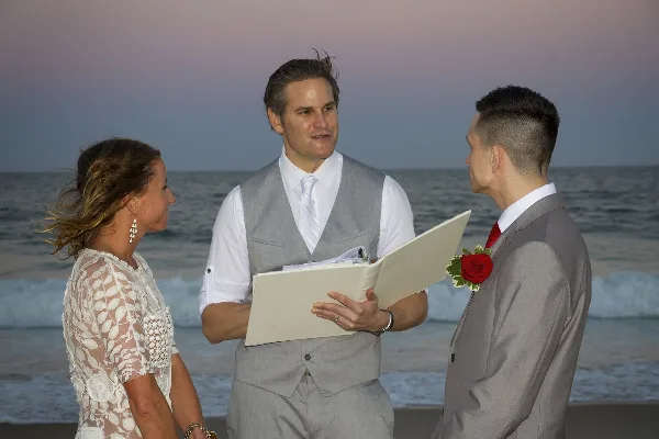 A wedding officiant in a grey vest and white shirt stands on a beach holding a folder, officiating a ceremony between a bride and groom.