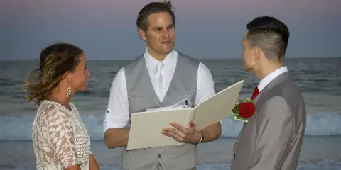 A wedding officiant in a grey vest and white shirt stands on a beach holding a folder, officiating a ceremony between a bride and groom.
