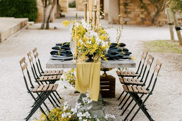 An outdoor event table set with wooden folding chairs, a yellow linen runner, and a floral centerpiece.