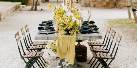 An outdoor event table set with wooden folding chairs, a yellow linen runner, and a floral centerpiece.