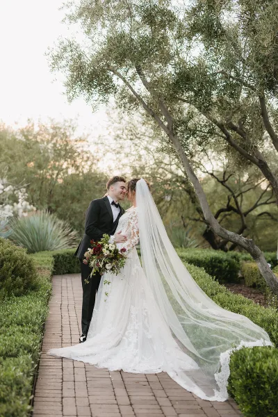 A bride and groom standing on a brick pathway in a garden, with the bride holding a bouquet and wearing a long veil.