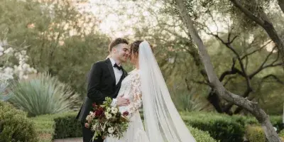 A bride and groom standing on a brick pathway in a garden, with the bride holding a bouquet and wearing a long veil.