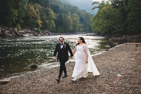 A bride and groom walking hand-in-hand along a rocky riverbank with a forested hillside in the background.