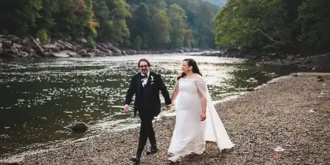 A bride and groom walking hand-in-hand along a rocky riverbank with a forested hillside in the background.