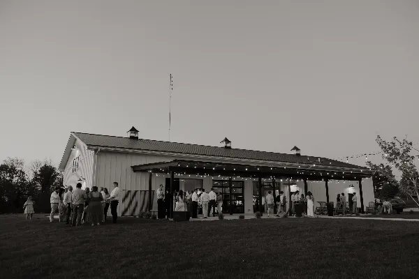 Exterior view of a white barn-style wedding venue with a covered patio and string lights, with guests gathered outside.