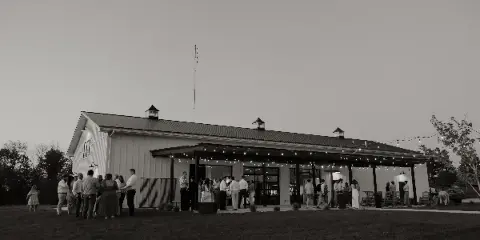 Exterior view of a white barn-style wedding venue with a covered patio and string lights, with guests gathered outside.