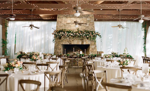 Interior of a wedding venue featuring a large stone fireplace, string lights, and round tables set with white linens and floral centerpieces.