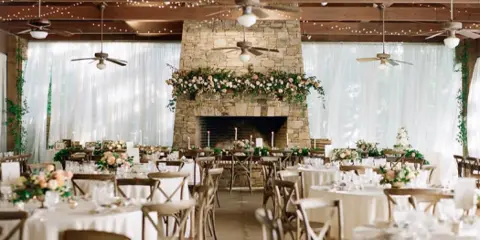 Interior of a wedding venue featuring a large stone fireplace, string lights, and round tables set with white linens and floral centerpieces.