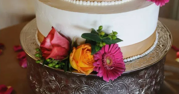 A close-up of a white and gold tiered cake decorated with red, orange, and pink flowers on an ornate silver stand.