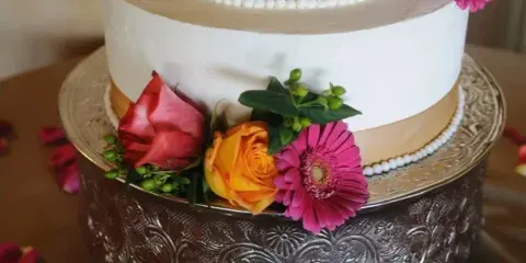 A close-up of a white and gold tiered cake decorated with red, orange, and pink flowers on an ornate silver stand.