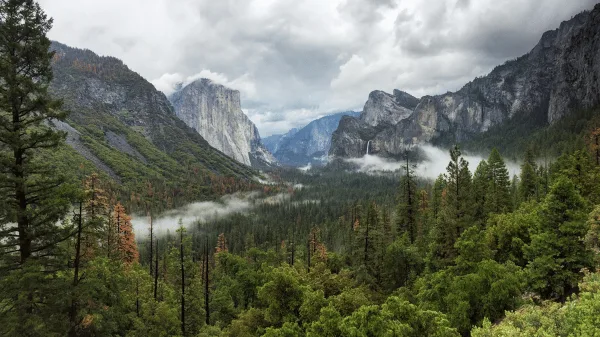 A scenic view of a mountain valley with a waterfall, surrounded by dense evergreen forests and rocky peaks under a cloudy sky.