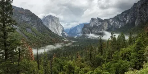 A scenic view of a mountain valley with a waterfall, surrounded by dense evergreen forests and rocky peaks under a cloudy sky.