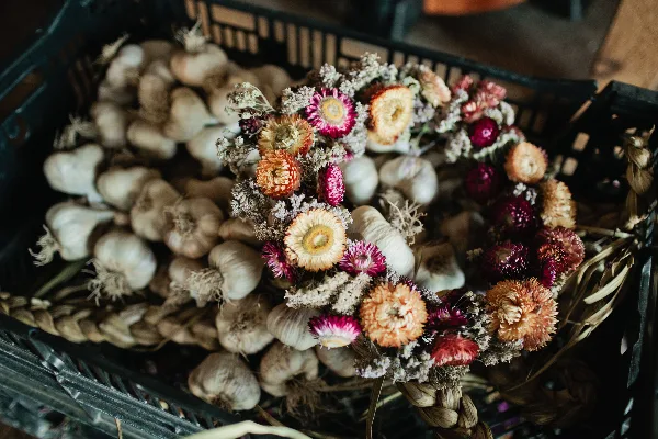 A dried flower crown with orange and pink blooms resting on a black crate filled with garlic bulbs.