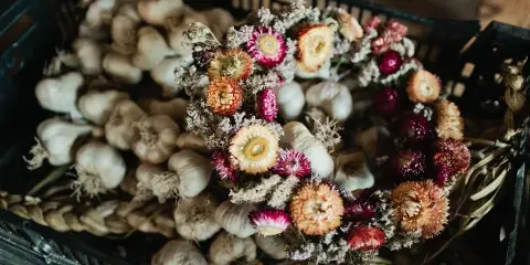 A dried flower crown with orange and pink blooms resting on a black crate filled with garlic bulbs.