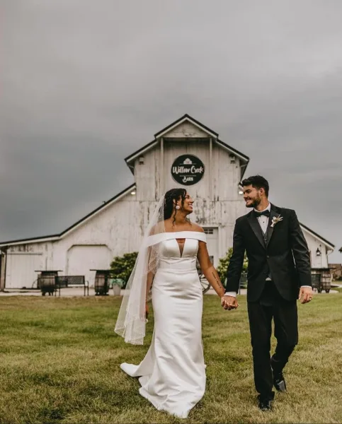 A bride and groom walking hand-in-hand on a lawn in front of a white historic barn with a 'Willow Creek Barn' sign.