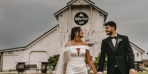 A bride and groom walking hand-in-hand on a lawn in front of a white historic barn with a 'Willow Creek Barn' sign.