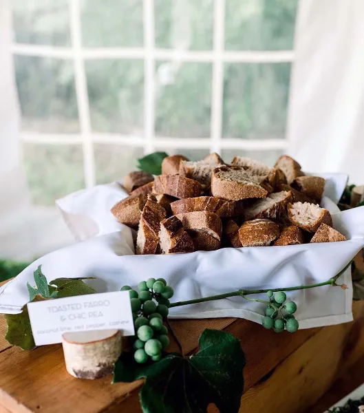 A white cloth-lined basket filled with rustic bread pieces, garnished with green grapes and leaves, accompanied by a small menu card.