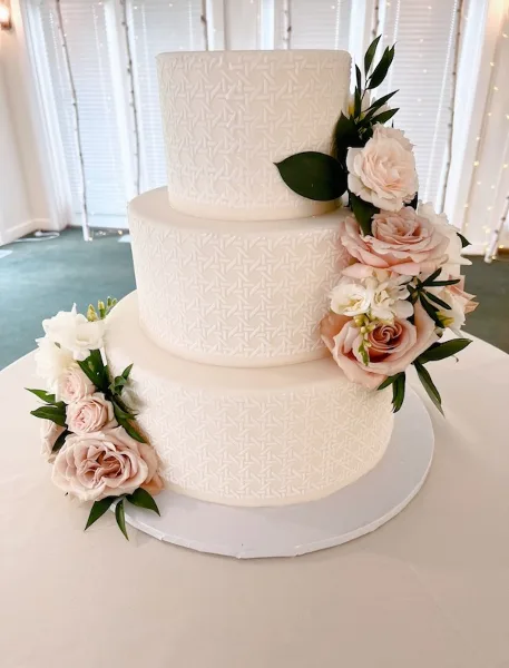 Three-tier white wedding cake with patterned fondant and cascading pink and white roses displayed on a white table.