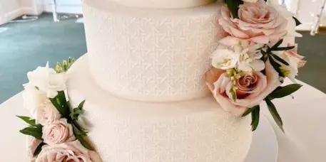 Three-tier white wedding cake with patterned fondant and cascading pink and white roses displayed on a white table.