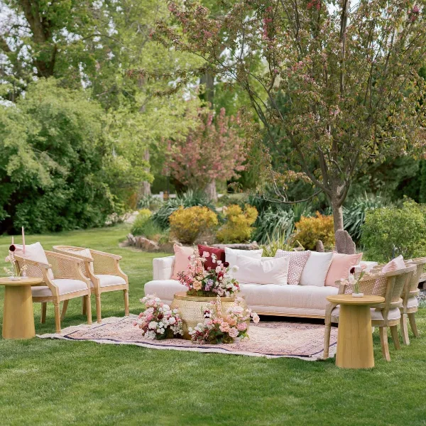 An outdoor garden lounge set featuring a cream sofa, cane armchairs, and side tables arranged on a patterned rug.