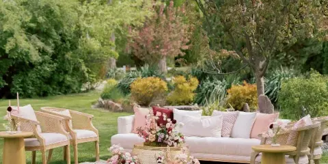An outdoor garden lounge set featuring a cream sofa, cane armchairs, and side tables arranged on a patterned rug.
