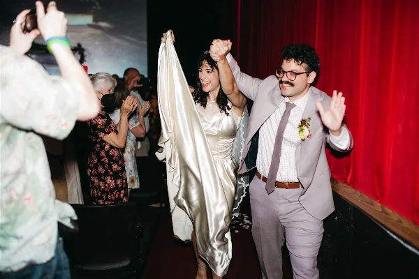 A bride and groom holding hands and walking down an aisle in a theater, captured in a candid, documentary style.