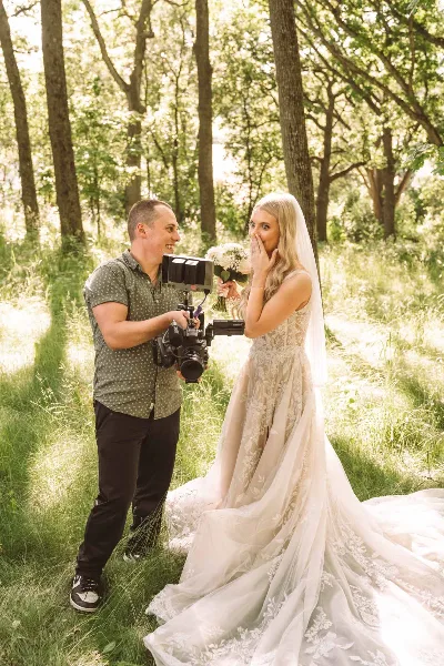 A videographer filming a bride in a wedding dress in a wooded area.