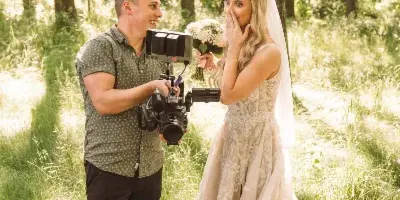 A videographer filming a bride in a wedding dress in a wooded area.