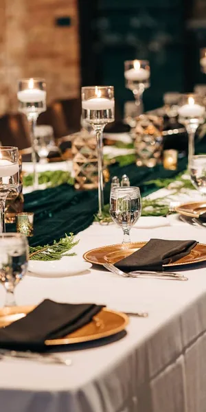 A formal event table setting featuring gold-rimmed plates, black napkins, tall candle holders, and greenery on a white tablecloth.