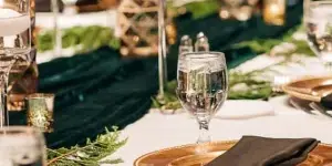 A formal event table setting featuring gold-rimmed plates, black napkins, tall candle holders, and greenery on a white tablecloth.