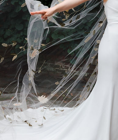 A bride in a white wedding dress holding up an embroidered sheer veil.