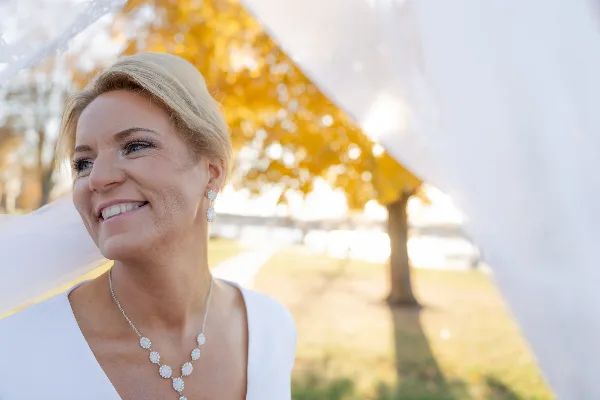 A smiling blonde bride wearing a white dress and necklace, photographed outdoors with a soft-focus background of autumn trees.