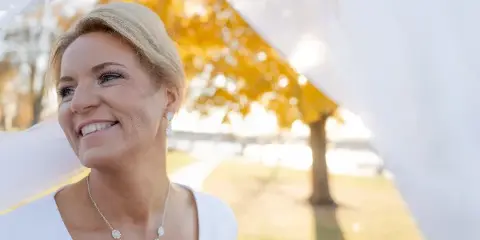 A smiling blonde bride wearing a white dress and necklace, photographed outdoors with a soft-focus background of autumn trees.