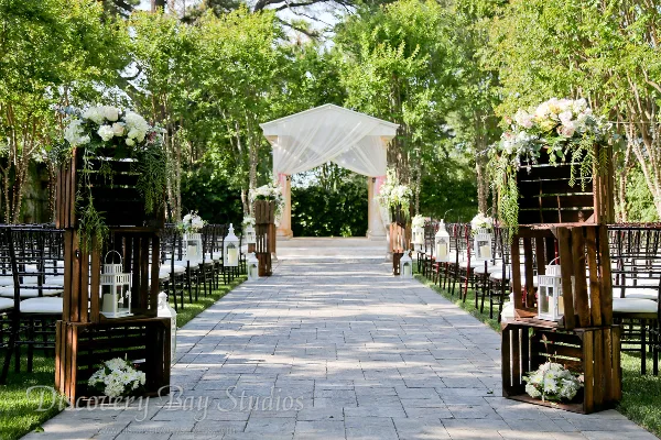 Outdoor garden wedding aisle with stone pavers, floral arrangements, and a white draped archway.