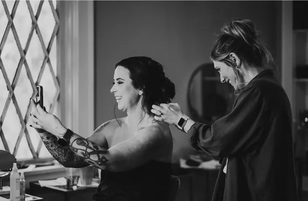 A black and white photo of a hairstylist styling a bride's hair while the bride takes a selfie.